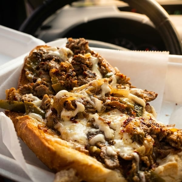Cheesesteak sandwich with melted cheese, steak, and peppers in a white takeout container inside a car.